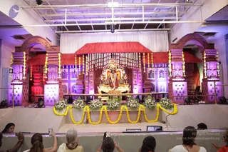 A photograph of a decorated indoor stage set for a ceremony, lit with purple and red lights. Two ornate arched pillars in terracotta and pink frame the stage, and a red-roofed canopy with white drapes hangs above a golden throne-like seat at the centre holding a deity murti or seated figure adorned with flower garlands. Yellow marigold garlands drape the stage edge over white flower arrangements. Several seated devotees in the foreground, including a woman in white and a woman with grey hair, some raising phones to photograph the stage. The seated central figure is a statue or decorated murti rather than a clearly visible living person.