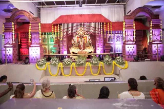 Photograph of a decorated stage set as a temple-style shrine with pink-red arched gateways, a red-tiled roof, and purple-lit pillars. At the centre sits a golden throne with a large deity murti or figure adorned with garlands and flanked by lions. Yellow marigold garlands hang across the stage and along the front railing, with white flower arrangements below. Viewers seated in the foreground include women in white and cream, one with a dark garland, and a woman in orange at the right edge, several raising mobile phones to photograph the stage. A small monitor sits on the railing.