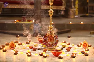 A brass lamp stand with a tall stem and a decorated base featuring a Ganesha motif and red, gold and green painted panels stands at the centre of a rangoli floor design dotted with yellow marigold petals. Many small clay diyas with lit flames surround it, along with red and orange clay bowls, and incense smoke rises beside the stand. No people are visible; the scene suggests a Diwali lamp display in a temple hall.