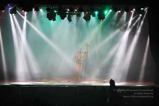 A stage performance under bright stage lights and haze, with green and white beams fanning upward. A lone figure stands centre stage holding a tall trident, dressed in a reddish-orange garment with a crown-like headpiece, presented as a costumed statue-like performer in the Shiva style. In the foreground at the bottom right, a camera operator and dark silhouettes of audience members are visible.