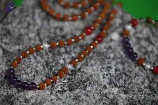 Close-up photograph of several beaded mala strands resting on a grey speckled granite surface against a green background. The strands mix brown rudraksha beads, small silver spacers, purple amethyst-coloured beads, red beads and white pearl-like beads. No people or portraits are visible in the frame.