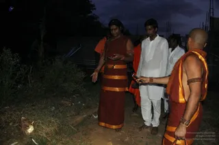 Photograph taken at dusk on rough ground beside overgrown shrubs and a construction site with bamboo scaffolding. A man with long dark hair, wearing a maroon robe with broad gold borders, rudraksha mala and orange-bangled arm, holds a mobile phone and gestures with his open hand, possibly a person by his long hair, saffron-gold ceremonial dress and rudraksha cues. Beside him a man in white kurta and white dhoti stands barefoot, and a shaved-headed attendant in saffron and gold-bordered cloth holds a phone out toward the group. Two more men in white stand behind them.