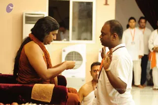 Photograph of a satsang-style exchange in a cream-walled room with white air coolers. A woman with long dark hair, wearing a rust-orange sleeveless garment and a rudraksha mala, sits on a maroon cushioned seat and holds a small object toward a man in a white shirt with an orange lanyard and a rudraksha mala, who stands before her with palms raised in greeting. Behind them a seated man in a white dhoti with a red forehead mark sits low, and two men in white kurtas with orange stoles stand near a curtain. The woman's appearance is not sufficient to confirm the a person candidate; she is described by role only, as a seated speaker in orange robes.