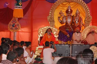 A seated bearded man in orange robes with a saffron-orange shawl and a marigold garland sits on an ornate gold throne speaking into a microphone under an orange tent. Behind him stands a large gilded frame holding two painted deity murti: the figure on the left has a red crown, multiple arms and a trident-like staff, and wears a garland and purple skirt, while the figure on the right wears a crown and red garland. To the right, a bearded man in a white kurta with a white turban stands beside a seated bearded figure in white. In the foreground, devotees sit on the floor, some in white dhotis, and a cameraman in white films the stage above two monitors. a person seated on the throne is the central speaker.