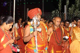Night procession photograph with a central figure in an orange-red turban and orange-red robe with a gold-coloured layered necklace and a flower garland in yellow and blue, holding a silver trident (trishul) on a long silver staff, smiling. Beside him a bald man in orange-red robes with a rudraksha mala and a brown shawl holds a camera. Around them devotees in orange and saffron garments, some with white forehead markings, a woman in an orange-yellow sari at right, a woman at left holding a staff and a phone. String lights and green plants form the background behind a mesh fence. a person: the central figure wears an orange turban, rudraksha-style layered gold ornaments and a ceremonial garland, with a trishul.
