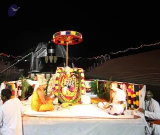 Night-time outdoor ritual at a temple-like setting with a large dark kalasha-shaped structure and a tall pillar behind. A deity murti is decorated with marigold and flower garlands and placed on a raised white-draped platform framed by green leaves and colourful garland strands. A man in an orange robe and a turban-like head covering sits cross-legged at the left of the platform, facing the murti and making an offering. Beside him, a second man in white clothing sits seated and tends the offerings. A young man in white stands at the far right edge, and a fourth figure is partly visible at the left. A multicoloured parasol canopy is raised above the deity. Strings of coloured lights run along the background roof line.