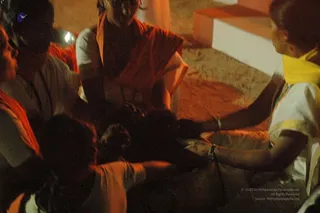 Dim night-time photograph of a small group seated on the ground near a sandy floor and a stepped platform. In the foreground, a woman in a white sari with a yellow border and yellow shoulder cloth reaches forward with bangled arms, hands together over an object held low between the group. Behind her, a person in an orange-red wrap sits upright beside another person in white, with a figure in a dark top at the left edge. Several people are partly visible, cropped at the edges. Activity suggests a ritual preparation, possibly the Navapashanam offering for consecrating a Linga, though the object itself is not clearly visible. Soft warm lighting dominates the scene.