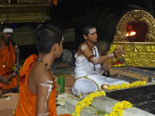 Night puja at an outdoor shrine decorated with yellow marigold garlands. A young priest in white dhoti and shawl, with white tilak and rudraksha malas, sits before a gilded shrine holding an oil lamp flame while ringing a handbell. Two shaven-headed young men in saffron robes sit nearby, one singing into a microphone with a white headband, the other with his back to the camera showing white body markings. A gold palanquin-like structure stands behind them, with brass vessels and a kalasha-style pot in the foreground. Text overlay at the lower right is mostly obscured.