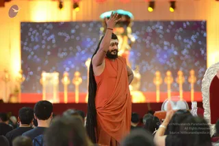 A photograph from a large hall event showing a man standing on a stage in an orange-saffron draped garment, long dark hair falling down his back, a white tilak on his forehead, and his right arm raised in a greeting gesture. Behind him, a large glowing backdrop with a starry bokeh pattern shows a golden deity murti and rows of golden lamps. Audience members in dark clothes sit in the foreground, one raising a hand. Swamiji (probable): the figure has long swept-back hair, saffron clothing and a tilak, matching cues for the page's named teacher.