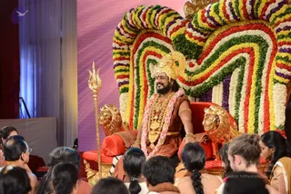 A man seated on a red and gold throne with lion-head armrests, in front of a large arch of multicoloured flower garlands in red, yellow, green, white and purple. He wears a gold crown, a gold necklace, a long pink and white flower garland, gold bangles and a rust-orange draped garment, and holds a golden trident staff at his side. Seated devotees in the foreground, several with hair tied back, face the stage.