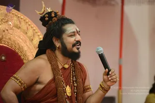 A bearded man with long dreadlocked hair wound into a topknot adorned with rudraksha beads and a gold crescent ornament speaks into a black handheld microphone. He wears a rust-orange wrap over bare shoulders, heavy brown rudraksha malas, a gold pendant necklace, gold armlets and bangles, and a white-and-red forehead mark. Behind him is an ornate gold and red throne, and the pink backdrop suggests a seated discourse setting.