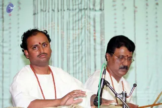 Two men in white ritual-style clothes perform music on a stage with a pale green draped backdrop hung with strands of beads. The man on the left, with curly hair and a red bindi, wears a red rudraksha mala and plays a drum with his hands. Beside him, a man in glasses and a red bindi, also wearing a red rudraksha mala, plays another drum. A microphone on a stand with a green cable sits between them.