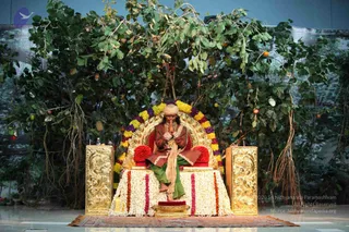 A seated figure sits on an ornate gold throne decorated with a marigold and jasmine garland, his palms pressed together in anjali. He wears a turban, a rust-brown jacket and a green wrap, with a cream shawl draped over his shoulders. The throne stands on a white flower-covered dais before a backdrop of leafy branches hung with fruit. A small brass offering dish sits at the base of the dais and a red carpet lies below. a person appearance cues are present but partial, so identity is uncertain.