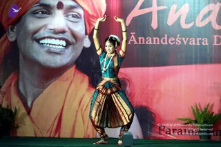 A female dancer performs on a stage in a green and gold Bharatanatyam costume with a gold-bordered skirt, a green blouse, a jewelled necklace and a white flower adornment in her hair. Her arms are raised above her head in a classical pose, and she wears ankle bells and sandals. Behind her is a large backdrop showing a smiling man in a saffron-orange turban and robes with a red tilak on his forehead, alongside large printed text reading "Ananda" and "Ānandeśvara D..." plus the partial word "Parama..." on the red panel. Potted plants flank the stage. A cultural performance is taking place at the event.