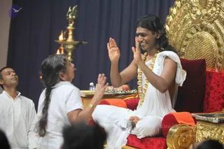 A seated man on a gilded throne with red cushions smiles and raises both palms toward a woman with a long grey braid in a white top who faces him with her hands raised. He wears a white dhoti and a white shawl over one shoulder, a gold-bead necklace, and a red tilak on his forehead. A brass oil lamp with two lit flames stands behind them against a dark blue curtain. A man in white kurta sits at left with his eyes lifted upward, and blurred heads of audience members appear in the foreground at the bottom edge.