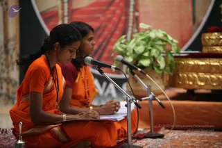 Two women seated cross-legged on a red and orange patterned rug recite from printed booklets at microphones during a satsang. Both wear orange saris and one wears a maroon top beneath her orange sari; each has a rudraksha-style mala. A small bell sits on the rug in the foreground, a potted green plant and a gold-coloured ceremonial box with red cushion are to the right, and a large red backdrop with partial lettering is behind them. No a person is visible in this image.