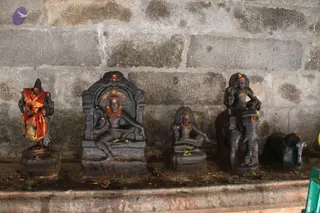 A photograph of four dark stone murtis set in a row against a weathered grey brick wall, with scattered flower petals and leaves on the ledge below. From left, a standing figure wears an orange and green striped cloth draped over the shoulders; a seated deity with a halo-like arch and red kumkum marks sits within a carved niche; a smaller seated figure sits on a low pedestal; and at right a figure is mounted on a bull-like animal beside a crouching animal carving. Red kumkum marks and vibhuti-like dots are visible on several figures. No living people are present.