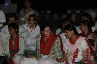 A nighttime group gathering of devotees seated on the ground, most in white or patterned clothing. Several women wear red shawls printed with white lettering, and one wears an orange beaded necklace. A woman in a green floral-patterned kurta and glasses smiles at the left, while a woman in a white floral top and pink dupatta gestures with her hand at right. A man in white sits behind them, and a woman in a white kurta with a braided hairstyle sits near the centre. Behind them a woman in a light sari with an orange necklace stands beside a striped wall.