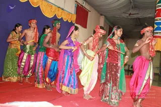A photograph of a group of young dancers performing on a stage with a red carpet and a blue draped backdrop with a yellow scalloped valance. Seven performers stand in a line holding short sticks, wearing colourful traditional costumes: bright pink, green, purple, blue and lime outfits with gold borders, and red and orange headcloths. Several boys are bare-chested with beaded necklaces, while the girls wear saris and matching jewellery and bangles. A ceiling fan and white tent-like ceiling are visible overhead.