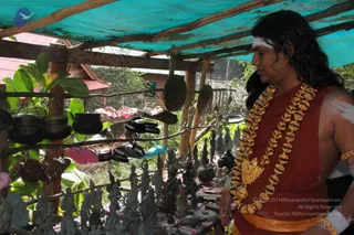A photograph of a man with long dark hair, wearing a rust-red wrap, a gold ceremonial necklace with a large pendant, gold armbands and a horizontal white forehead marking, standing under a blue tarpaulin at an open-air stall. He looks down at a table of small bronze deity figurines and brass bowls, while metal bells, bowls and sandals hang from wooden poles behind him. Greenery and a red-roofed building appear behind the stall.