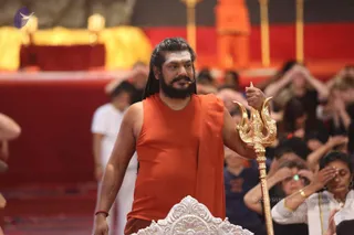A bearded man with long dark hair stands at a silver-edged ornate pedestal in a large red-draped hall, wearing a sleeveless saffron-orange garment with an orange shawl over one shoulder and a beaded bracelet on his wrist. He holds an ornate golden trident upright in one hand and gestures with the other. Behind him, a figure in orange stands on a raised platform with gold pillars. In the foreground and background, many seated participants in dark clothing rest their hands on their heads in a yoga posture, and a few people in white stand nearby. Probable Swamiji (long hair, saffron-orange clothing, trident and shawl) appears in person, performing a satsang-style address beside the throne-like pedestal.