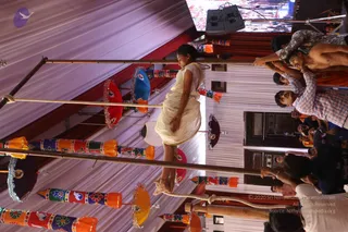 A photograph, rotated sideways, of a festival or temple-stage setup under a white draped canopy with red and orange decorative strips. A young woman in a cream-colored sari with a white border stands on a wooden bar or rope-walk structure, one hand touching a horizontal pole. Several colorful painted poles hang with multicolored tassel-like bunting, round red, blue and yellow umbrella-shaped decorations with silver tinsel. On the right, a crowd of men in shirts, some shirtless with orange cloth, reach up toward the pole structure. A speaker and screen are visible above.