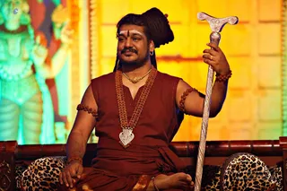 A man with long dark hair coiled on his head and a white and red tilak on his forehead sits on a leopard-print cushioned seat with a carved wooden frame. He wears a rust-red sleeveless garment with a wrapped waist cloth, multiple strands of rudraksha beads across his chest and arms, and a silver pendant with a gold chain. He holds a tall silver staff with a T-shaped handle in his right hand. Behind him, a green-lit deity murti stands against a golden backdrop. The figure has distinctive long swept-back hair, rudraksha malas and a red ascetic robe, so this is an appearance-based a person in person, seated in a ceremonial setting.