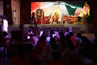Indoor hall with an audience seated in rows facing a stage backed by a large projected or painted backdrop of a red-toned temple tower gopuram, a mountain and a starry sky. On the stage a seated figure in saffron-orange robes sits before a lit temple-like shrine decorated with white and gold, with a white statue-like figure beside him and a garlanded deity figure at the right edge. A tripod-mounted camera operator stands at the left in white clothing. Audience members wear mostly orange, red and white clothing, viewed from behind.