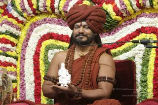 A seated man in an orange-red turban and orange-red robe holds a small white statue of a seated figure in his open palms. He wears layered rudraksha malas, rudraksha bracelets on both upper arms, and a gold pendant necklace, with a red tilak on his forehead. He sits on a dark red velvet throne before a microphone stand, with a backdrop of concentric arcs of red, yellow, white, and purple flower garlands. Behind him at left is a brass vessel. The setting suggests a satsang gathering. Swamiji (probable): the man has long swept-back hair, a turban, saffron-orange clothing, and rudraksha malas, consistent with the page's reference to Nithyananda Paramashivam.