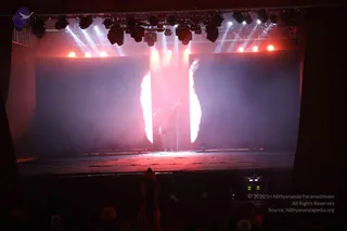 A stage performance in a dark hall with a large rear projection screen showing a pale orange flame-like glow and smoke. A lone figure stands near the centre of the stage in silhouette, lit from behind by warm light. Rows of stage lights hang above, and the audience is seen as dark shapes in the foreground, with a few hands raised at the bottom edge. The scene looks like a stage show or darshan presentation.