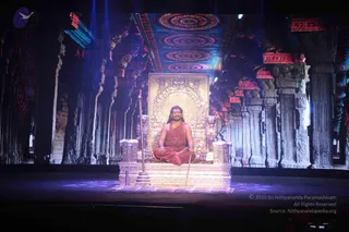 A seated figure on an ornate gold-and-silver throne at the centre of a large stage, flanked by carved lion-shaped armrests and a standing staff. The figure wears a saffron-orange robe, a red-brown rudraksha mala and long hair swept back, with legs crossed in a meditative pose. Behind the throne, a huge projected backdrop shows a temple hall with carved columns, ornate ceilings with concentric circular patterns and red-trimmed brackets. Purple stage lighting falls across the platform. No other people are visible in the frame.