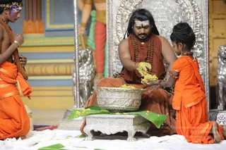 A seated man in an orange-red robe, with long dark hair, rudraksha beads and a white tripundra on his forehead, sits on an ornate silver throne with silver lion figures at its sides. He scoops yellow ritual powder from a large engraved silver bowl set on a silver stand, with a banana leaf below. A young girl in orange clothing kneels at right offering a handful of yellow substance, and a boy in orange with rudraksha beads kneels at left. The setting appears to be a ceremonial hall with painted columns behind.