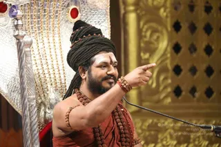 A photograph of a man in a rust-orange robe seated before an ornate silver and gold throne with red jewelled accents. He wears a tall dreadlocked turban, rudraksha malas across his chest and arms, and a white forehead with a central tilak. He points his right arm outward while speaking into a microphone on a stand, in front of a gold-carved wooden screen. a person: the rudraksha malas, long dreadlocks and ornate throne match cues associated with the presentation, and the page indicates his presence, so the in_person value applies.