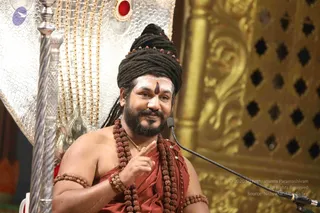 A man seated on an ornate throne speaks into a microphone during a satsang discourse, pointing upward with his right index finger. He wears a tall dark braided crown or turban, a white forehead marking with a red bindi, and a thick beard. Multiple rudraksha malas hang around his neck and wrists, and his body is draped in a red-orange cloth. Behind him stand a silver-and-gold throne backrest with scalloped panels and a gilded latticed wall with carved floral motifs. Swamiji (probable) appears in this photo as the seated speaker; the appearance cues are the crown, rudraksha malas and saffron-red garment, and the page identifies the event as a live satsang.