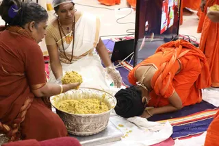 A photograph of a devotee in orange robes with long dreadlocked hair bowing forehead-down on a striped mat, a sign of deep prostration. Beside them, two women kneel and serve yellow rice from a large ornate silver bowl; the woman at center wears a white and gold sari, rudraksha beads, a gold pendant and latex gloves with a white hairnet, while the woman at left wears a rust-red sari and gold bangles. A laptop and a screen showing an orange-clad figure stand behind. Other orange-robed people are partially visible at the edges.