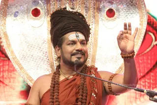 Photo of a man seated before an ornate silver-and-gold backdrop with red jewel-like medallions. He wears a tall dreadlocked headpiece topped with a crescent ornament, white horizontal tilak marks with a central dark line on his forehead, and a rudraksha mala with a gold chain. He is draped in a rust-orange cloth with bare shoulders and a rudraksha bracelet, smiling and raising his right hand while speaking into a microphone. Swamiji (probable): long swept-back dreadlocks, a crown-like crescent, saffron-orange clothing, rudraksha malas and a ceremonial gold necklace identify him as a presiding speaker at the event.