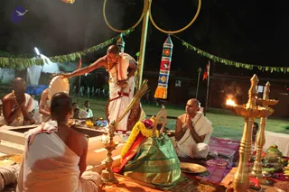 A night-time outdoor ritual photo. Several bald men in white dhotis and bare upper bodies sit on a raised platform around a fire pit. A standing priest in white dhoti and red-bordered cloth extends his hand over the fire, with gold necklaces on his chest. A seated devotee in white with palms joined faces the ceremony. Brass oil lamps with lit flames stand in the foreground, with fruit offerings, a green silk-bordered bundle with flower garlands, and a kalasha-like ritual setting. Gold rings hang from a pole overhead, with decorative banners and green leaf garlands strung across the scene.