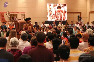 A seated man in a large brown leather armchair sits at the front of a hall facing a large audience. He wears a black turban-style headpiece, a saffron-orange robe with a gold-toned armband, and a long multi-strand necklace of beads and gold. His hands rest in front of him. Behind him on the left is a red fabric hanging, a trident leaning against the wall, and a framed religious painting. A large screen on the back wall shows a close-up of a figure in a black headpiece, red robe and bead necklaces standing at a microphone. Audience members in mostly white, orange and pastel clothing fill the room, many wearing lanyards, and a cameraman stands on the right.