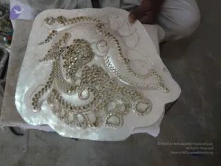 A close overhead photo of a craftsman's hands holding a large pale silver-white plaque on a low table. The plaque bears a raised, embossed floral and scroll pattern edged with rows of small round studs or rivets, with a leaf-like motif at the upper right. Only a partial arm in white clothing is visible at the top, with a bare hand resting on the plaque's edge. A concrete floor and a cloth-covered surface surround the work. No deity, ceremony or a person candidate is visible.