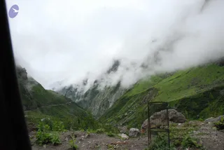 Photograph of a misty Himalayan valley seen through a vehicle window, with dark window frame at left. Low clouds drape steep green hillsides and rocky grey cliffs. A narrow winding road cuts along the slope at left, and boulders and scrubby green plants sit in the foreground. A rusted wire cage stands near a large rock at right. No people are visible.