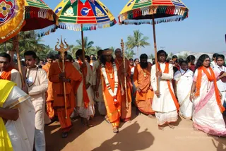 Outdoor procession on a sandy dirt road lined with palm trees under a clear blue sky. Men and women in saffron and orange shawls and robes walk behind multicoloured canopy umbrellas with fringed edges and floral motifs. At front left a man in orange holds a tall golden trident, and beside him another carries a golden staff. In the centre a bearded man in orange wears a crown-like headpiece, a long flower garland, and gold necklaces, appearing as a person in the ceremonial procession. Behind him a bearded man in orange and a man in white with an orange shawl follow, along with women in white saris with orange shawls and devotees in white.