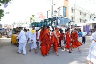 A street procession in a South Indian town moves past a white tourist bus and a yellow auto-rickshaw. At the centre, a bare-headed man in saffron cloth and a garland of orange beads walks barefoot beside a man in a red-orange turban and orange dhoti who carries a long staff. Behind them, several devotees in saffron and red robes follow, while men in white dhotis and yellow shawls walk on the left. Shop fronts, a roadside fruit stall and a camera operator line the street.