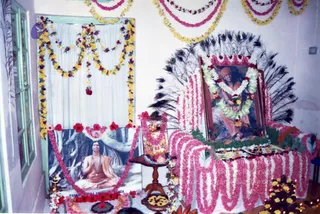 A photograph of a room decorated for Diwali with garlands of yellow and pink flowers hanging across the walls and a white drape. Two framed portraits sit on a flower-covered altar: on the left, a seated figure in saffron robes with rudraksha beads and hands in prayer, with a printed caption reading "PARAMAHAMSA NITHYANANDA" beneath it; on the right, a framed portrait of a saffron-clad figure seated in front of a rock backdrop, garlanded with white and red flowers, set before a fan of peacock feathers. Oil lamps burn at the altar base and a brass lamp and flower offerings lie on the table. No physical people are visible in the scene.