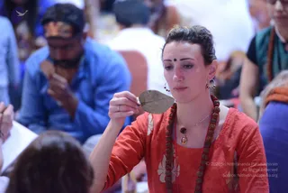 A woman in a red-orange embroidered kurta wears a long rudraksha mala and a pendant necklace, with a small bindi on her forehead. She holds a dry brown leaf up in her right hand and looks down at it, seated among other people in a crowded hall. Behind her a man in a blue shirt sits with his hand near his face. Blurred seated attendees fill the background.