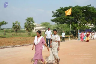 Outdoor photograph on a reddish-brown dirt courtyard. In the foreground two women walk side by side: the left one wears a pink salwar kameez with a white dupatta draped over her shoulder and sunglasses, and the right one wears a white saree with a brown patterned border and sandals. A saffron flag flies from a pole beside a tree and a street lamp. Behind them, a line of women in colourful sarees, including pink, yellow and red, and two men in white shirts walk across the grounds toward a multi-storey building with palm trees to the left. No deity, throne or a person figure is visible.