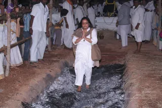 A woman in a white kurta and white trousers walks barefoot along a shallow trench of glowing embers and ash during a fire-walking ritual, her palms pressed together in anjali mudra and a brown shawl draped over her shoulders with a brown cloth bag on one arm. Behind a wooden barrier, a crowd of men in white dhotis and shirts stand with hands folded, and a decorated deity murti with garlands is visible in the background. The trench edges are lined with straw and earth.