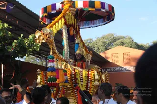 A golden ceremonial chariot procession at an orange-walled temple compound. A figure with long dark hair, a golden crown and an orange-saffron garment with a rudraksha-style necklace of beads sits on an ornate gold palanquin-style seat under a multicoloured fringed canopy in red, yellow, green, purple and blue. The seat and canopy are wrapped with marigold and yellow garlands and hanging tassels. Devotees in white and orange clothing surround the chariot, and a hand-held microphone is visible at lower centre. A signboard in Kannada script is on the orange wall behind. Trees and blue sky are in the background.
