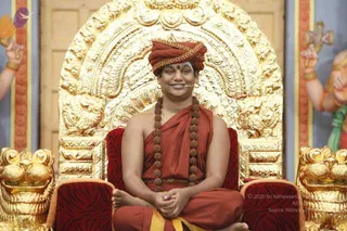 A man seated cross-legged on an ornate gilded throne with a large golden arched back and red velvet cushions with gold lion-head armrests, in a satsang setting. He wears a rust-red turban with a gold-braided band, a rust-red shawl and dhoti, a long rudraksha mala draped to his waist, and a tiny clip microphone at his cheek. He has a red bindi and holds his hands folded in his lap. Painted deity figures with raised hands and gold bangles flank the throne on both sides. Swamiji (probable) is shown in person with cues of a rust-red turban, saffron-toned shawl and rudraksha mala while seated on the throne.