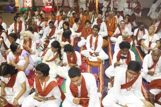 A large seated assembly of devotees in white clothing with orange-brown shawls fills a hall during a satsang session. Many wear orange-and-white lanyards and hold small bells or wooden clappers. A man near the centre wears a saffron/orange robe with a rudraksha-style necklace and sits on a striped mat, but his appearance is not clearly distinctive enough to confirm identity, so he is described only as a seated devotee. A deity murti with flower garlands is visible at the top left, beside a red-draped altar.