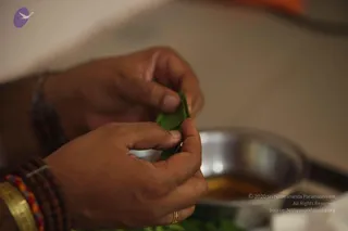 Close photograph of a man's hands folding or tearing a green leaf over a shallow stainless steel vessel holding a golden liquid. The wrists wear rudraksha-style beaded bracelets in brown and multicolour threads and a gold bangle; a gold ring is on one finger. The background is softly blurred, with a green leaf pile at the bottom edge.
